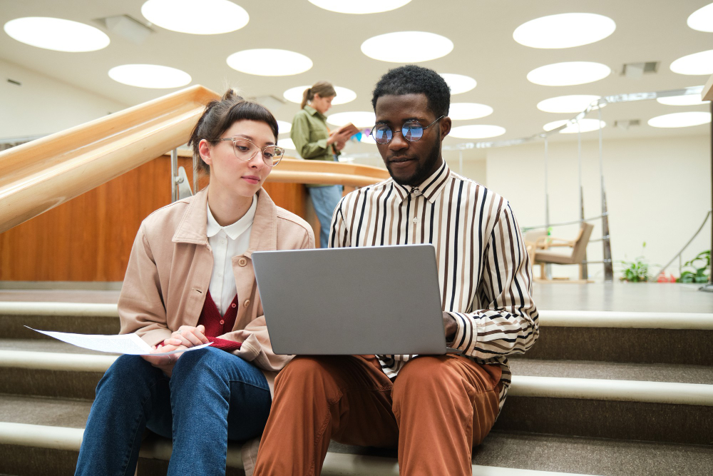african-man-using-laptop-studying-together-with-woman-while-they-sitting-stairs-library